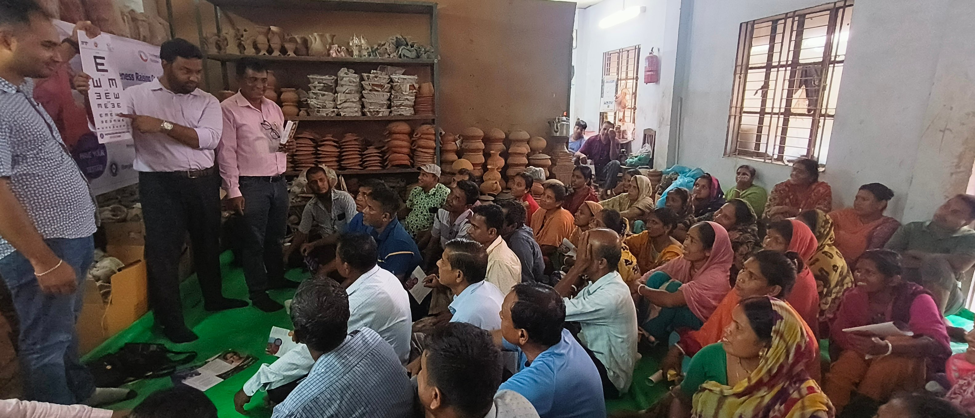 A group of Bangladeshi potters gathered closely together, listening to an eye health educator during a community awareness session in Bijoypur, Cumilla.
