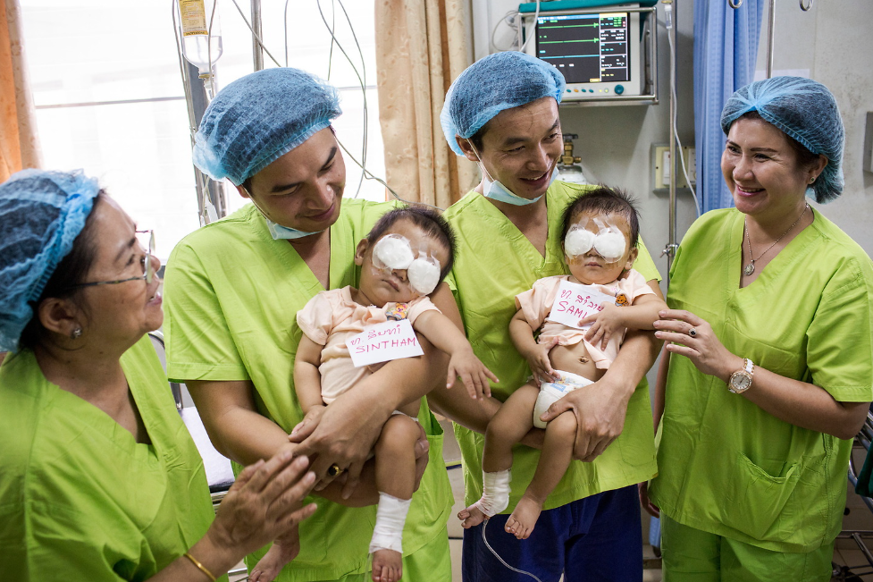 Seven-month-old twins Samlan and Sintham were born with cataracts and had not been able to see clearly. After travelling two days by boat for surgery, they are pictured with the surgical team, their eye patches still on, after their successful operations.