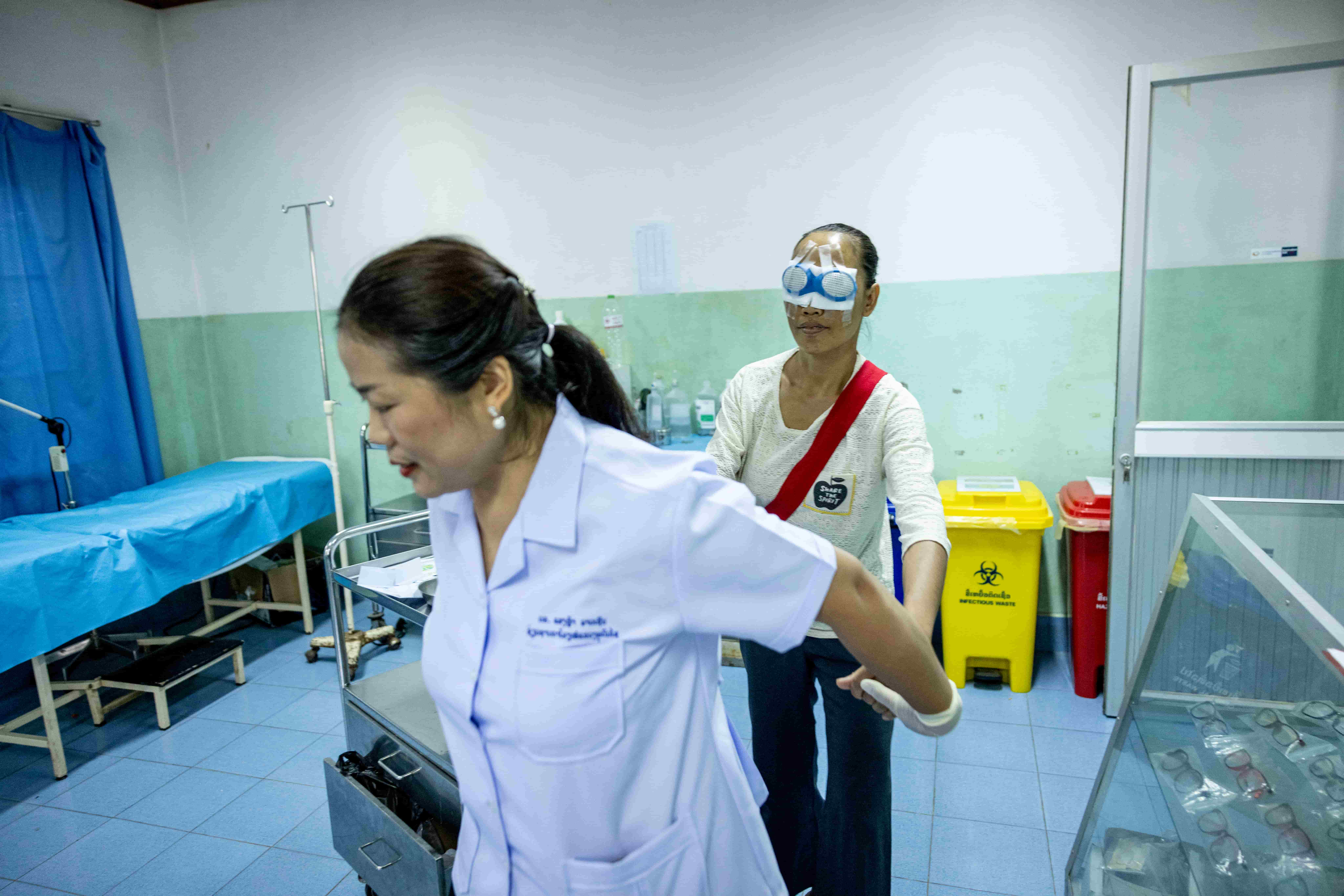 Dr Senglar walks in front, gently holding the hands of Lien, who has two eye patches after cataract surgery, guiding her safely from the surgical ward at Oudomxay Eye Hospital.