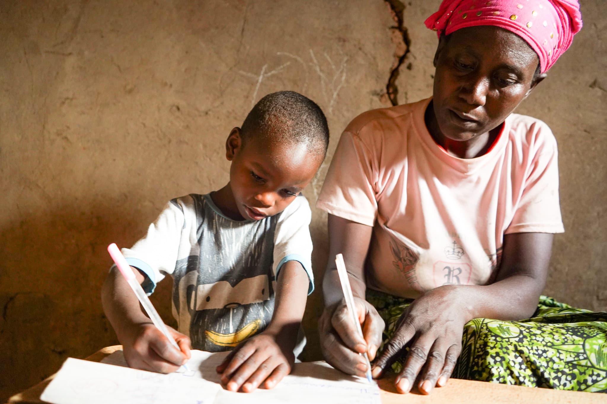 Blaise sits with his grandmother at home, focusing on his schoolwork as she supports him beside him, the two sharing a quiet moment of learning together.