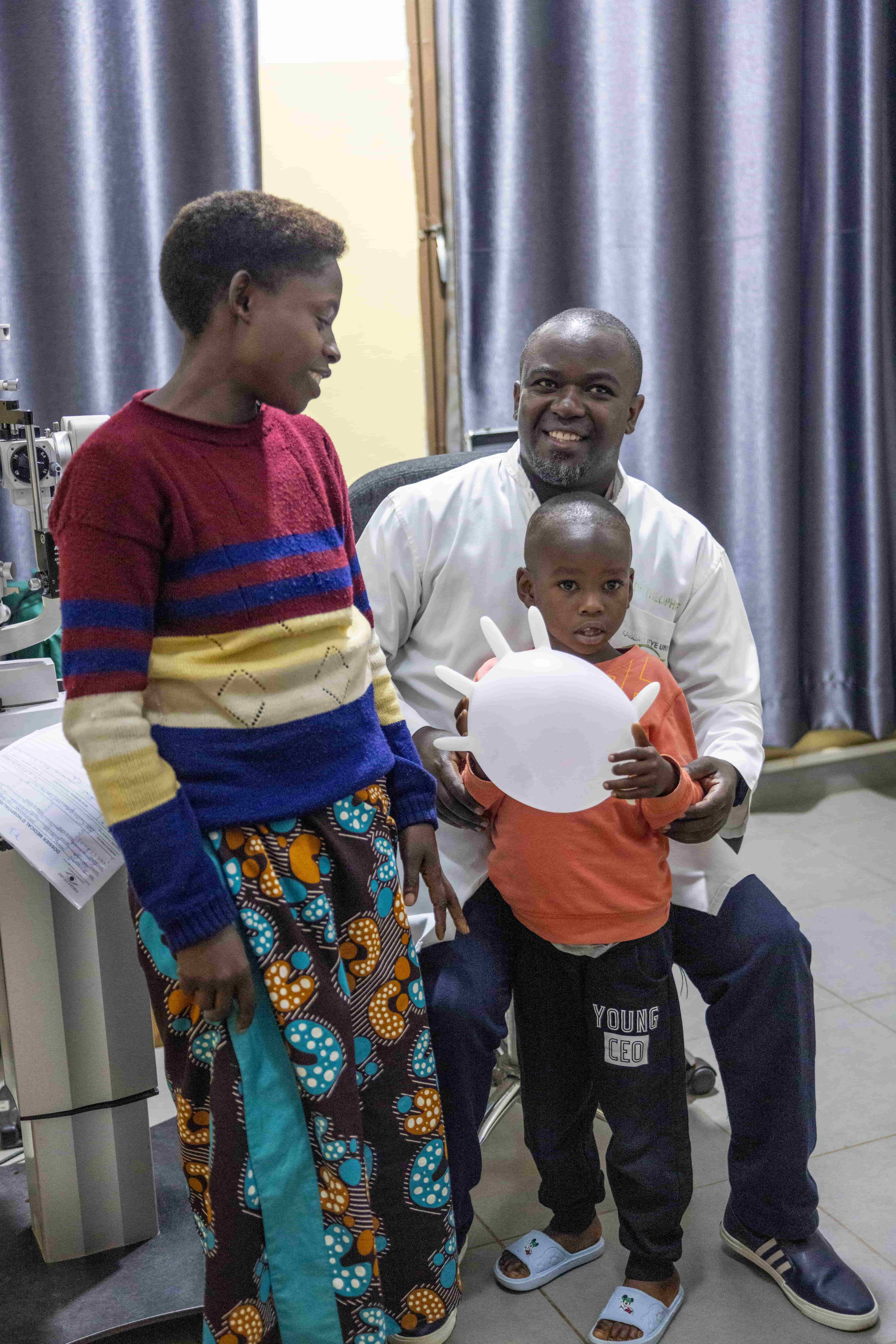 Blaise smiles as Dr Theophile inflates a surgical glove like a balloon to ease his nerves before surgery, while his mother Redempta stands close by, watching with reassurance.