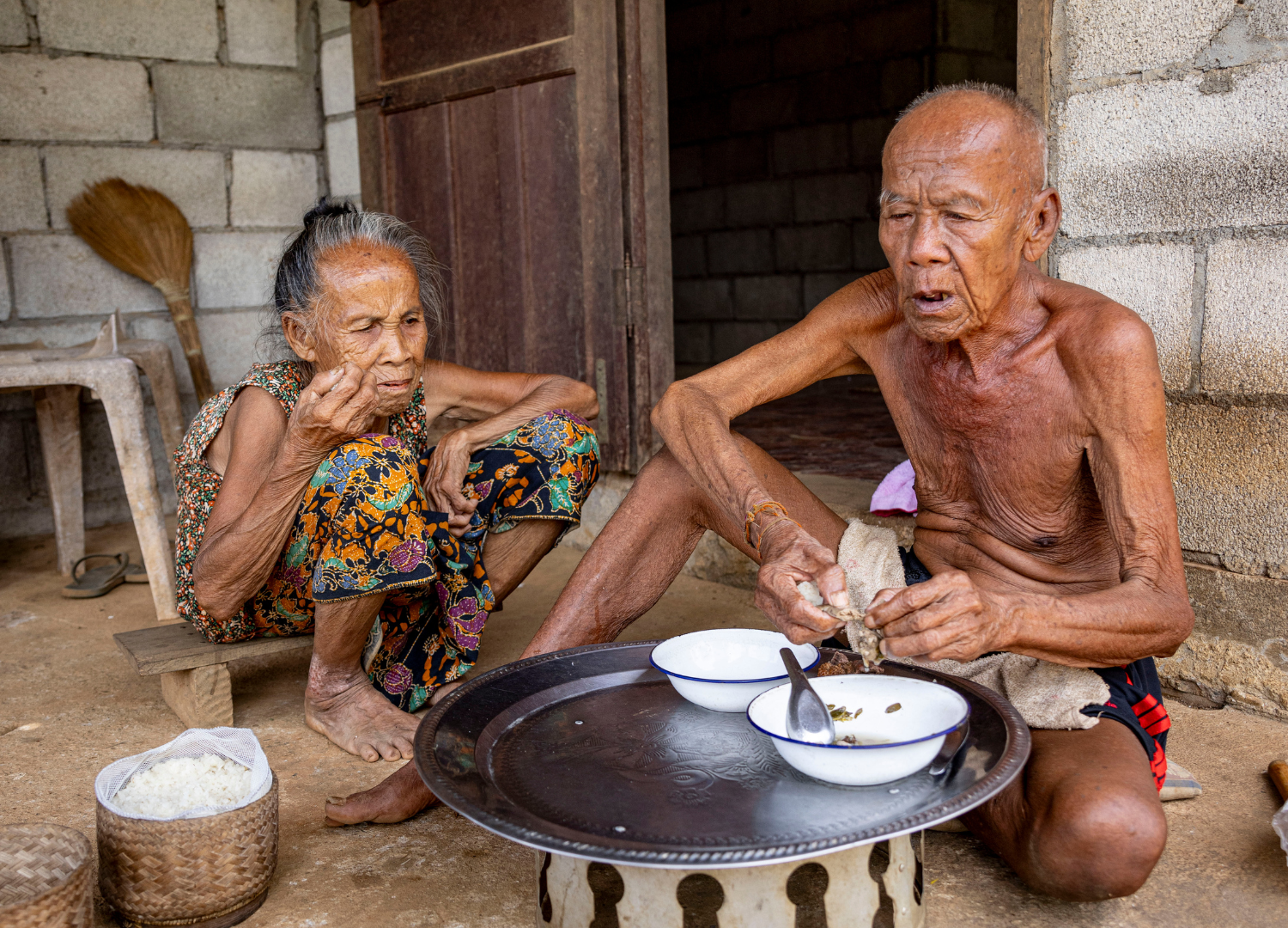 Thoung and Mai, crouching on the ground at their home to eat a meal.