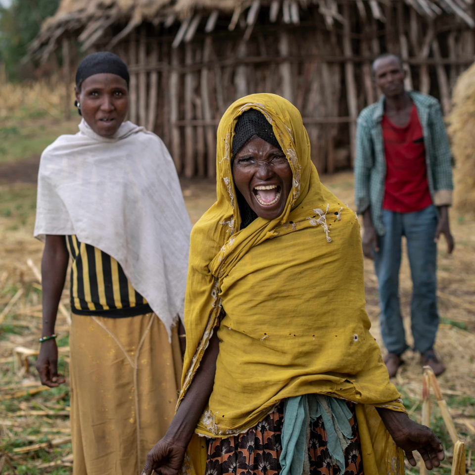 Hawiti wearing a yellow scarf, smiling in front of her mud brick home