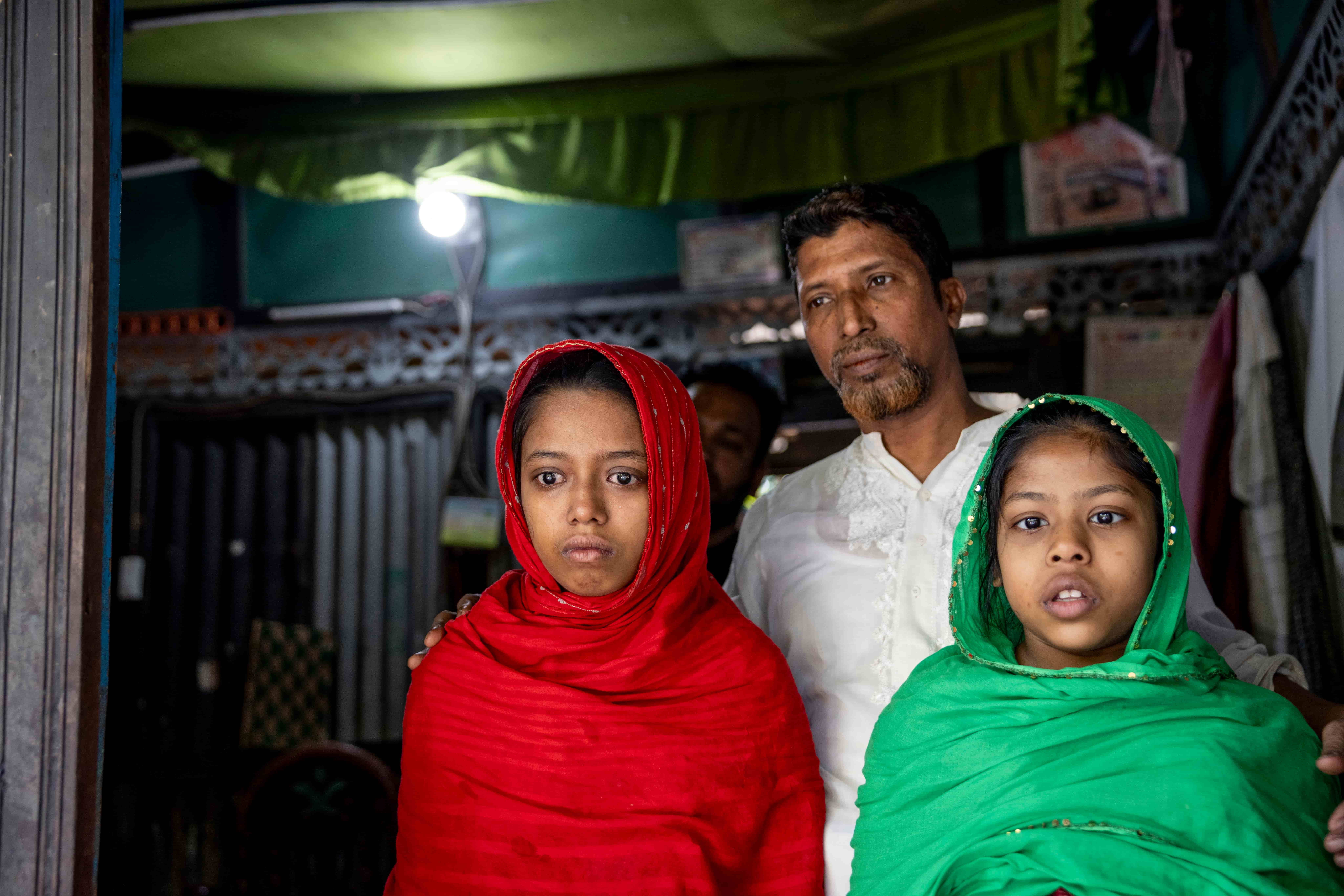Sumaiya and Habiba stand at home with their father, Md Awlad Hossain, positioned between them. He gently holds his daughters close inside their modest house in Chandpur, Bangladesh, as the sisters live with severe vision loss before surgery.