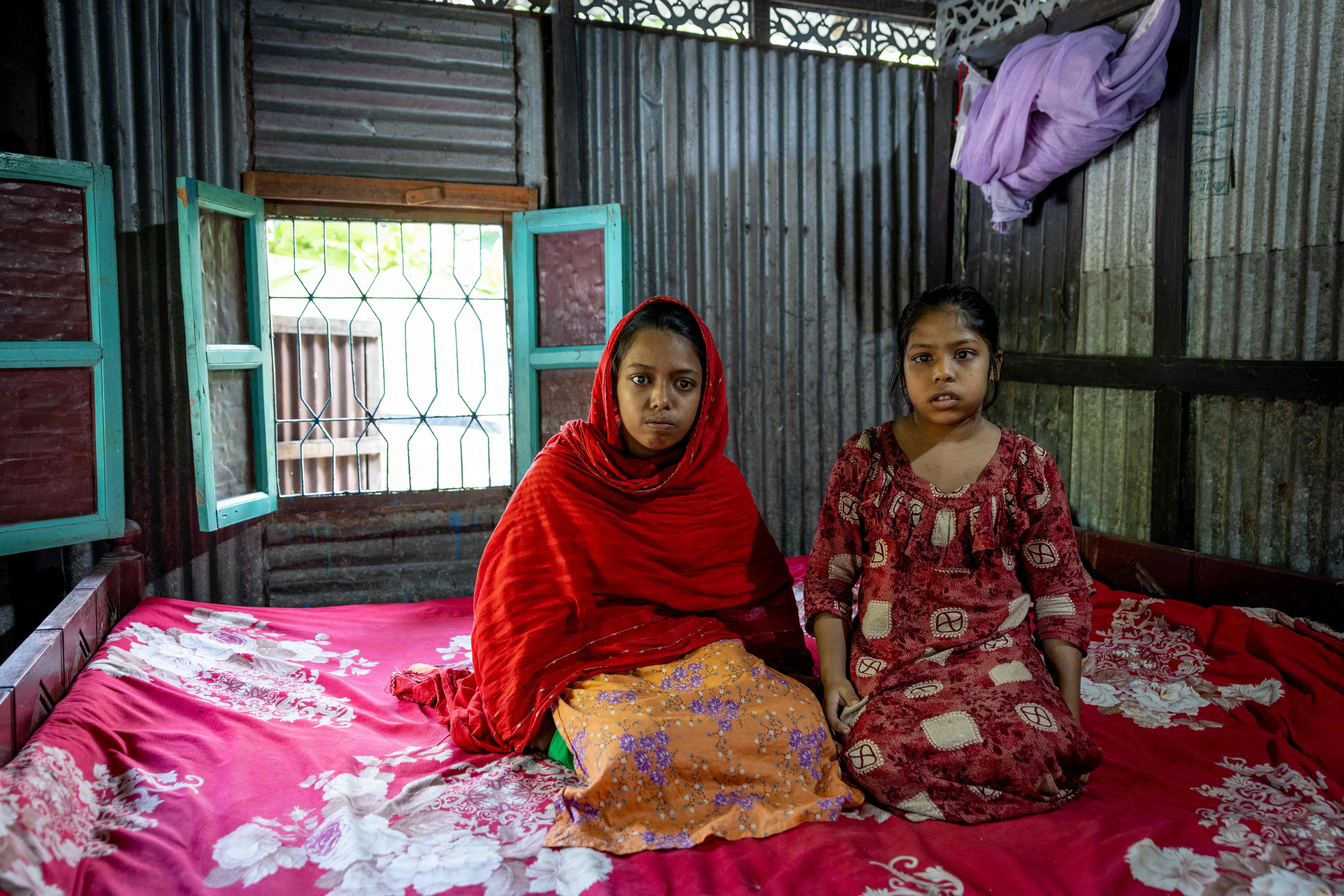 Sumaiya, 17, and Habiba, 14, sit side by side on a small bed inside their family home in Chandpur, Bangladesh. The sisters sit close together in the dimly lit room, their expressions quiet and uncertain as they live with severe vision loss before receiving cataract surgery.
