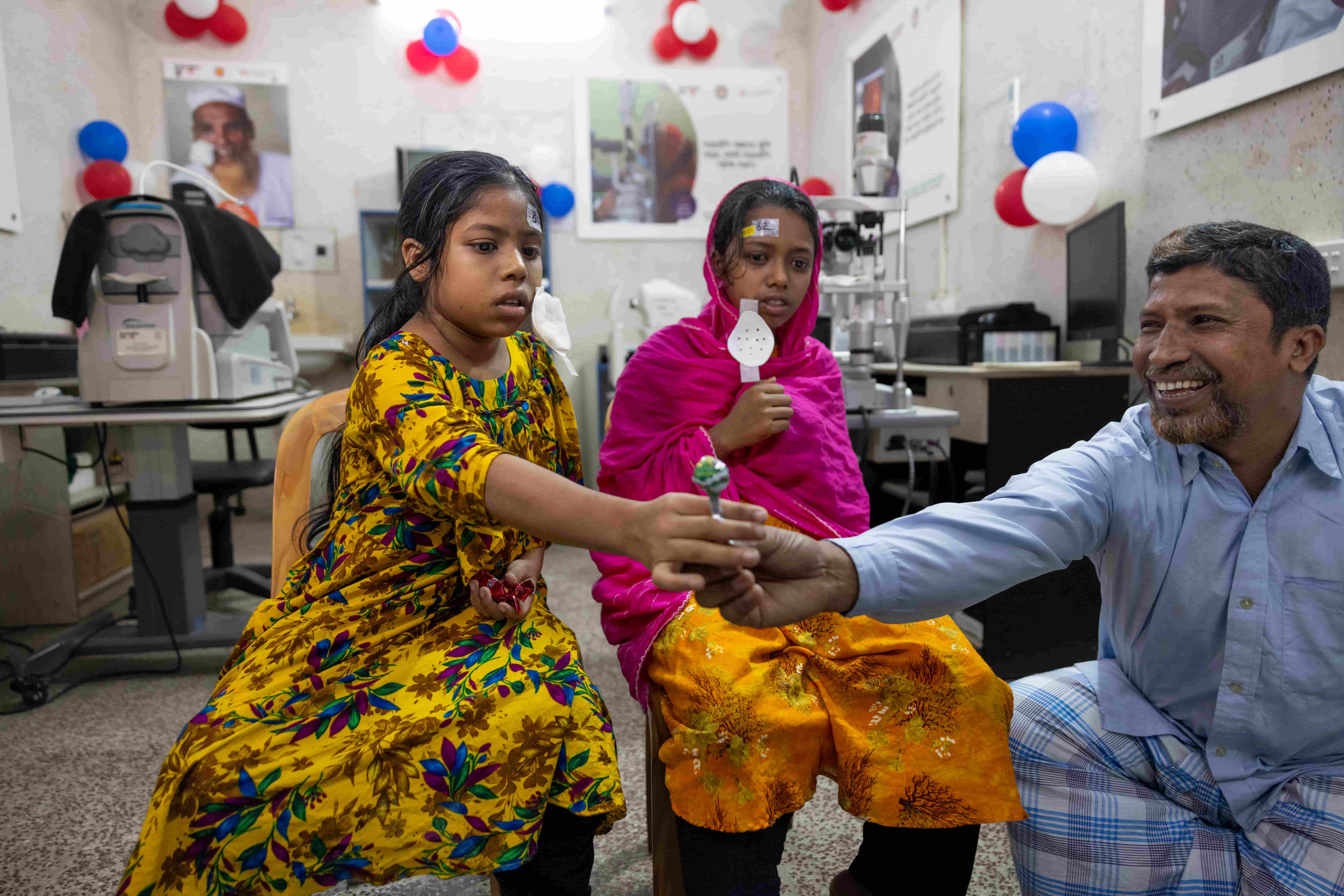 Habiba reaches confidently for a lollipop offered by a nurse as her eye patches are removed the day after surgery. Sumaiya sits nearby, while their parents, Hajera and Md Awlad Hossain, watch with tears of joy and relief, celebrating their daughters&rsquo; restored sight and new opportunities.