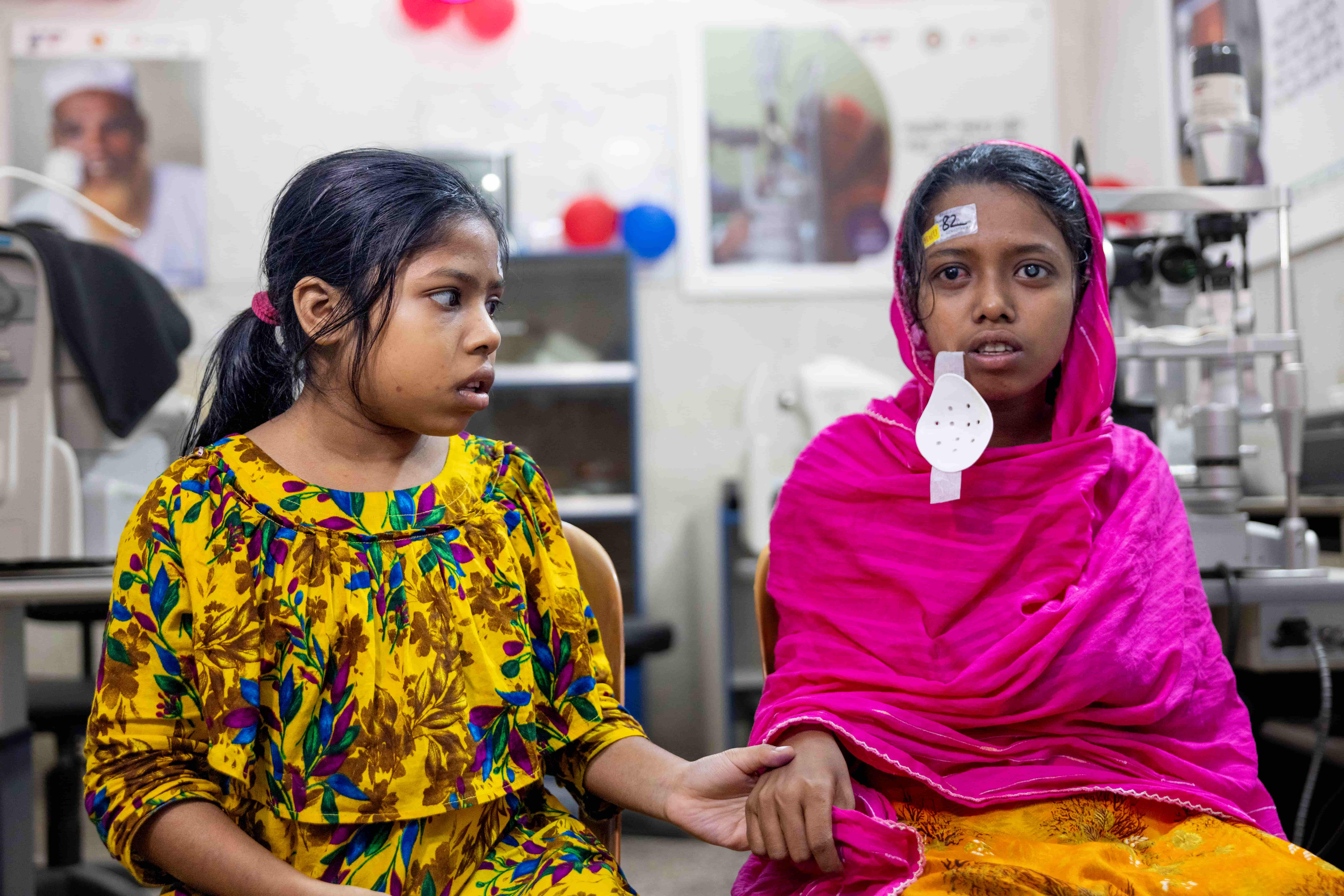 Sumaiya and Habiba sit side by side with their eye patches removed after surgery. They are looking forward, adjusting to their restored sight, with neutral expressions.