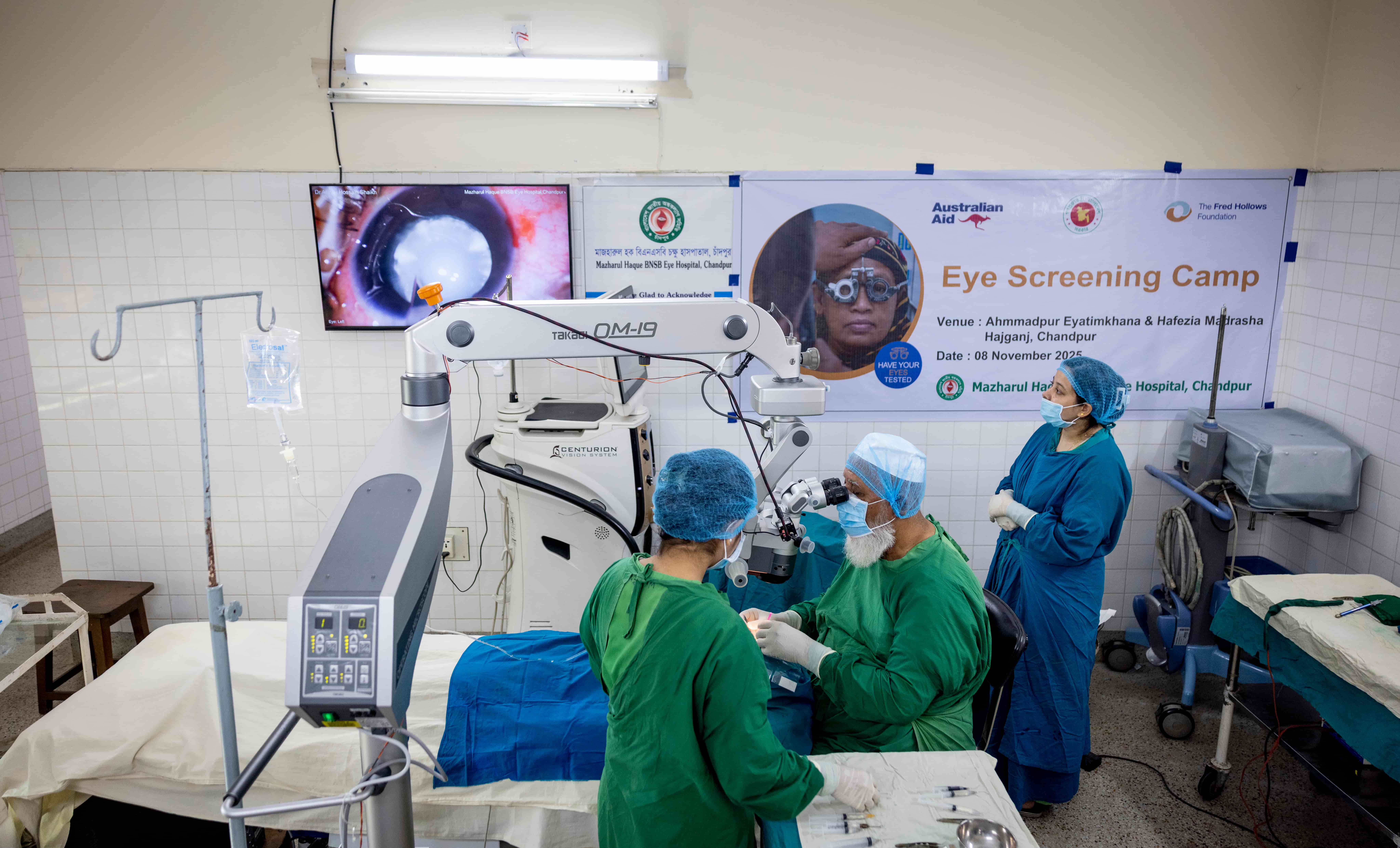 Inside the operating theatre at Mazharul Haque BNSB Hospital in Chandpur, a young girl undergoes cataract surgery while the surgical team works carefully under bright theatre lights. Medical equipment surrounds the operating table as staff focus intently on the procedure.