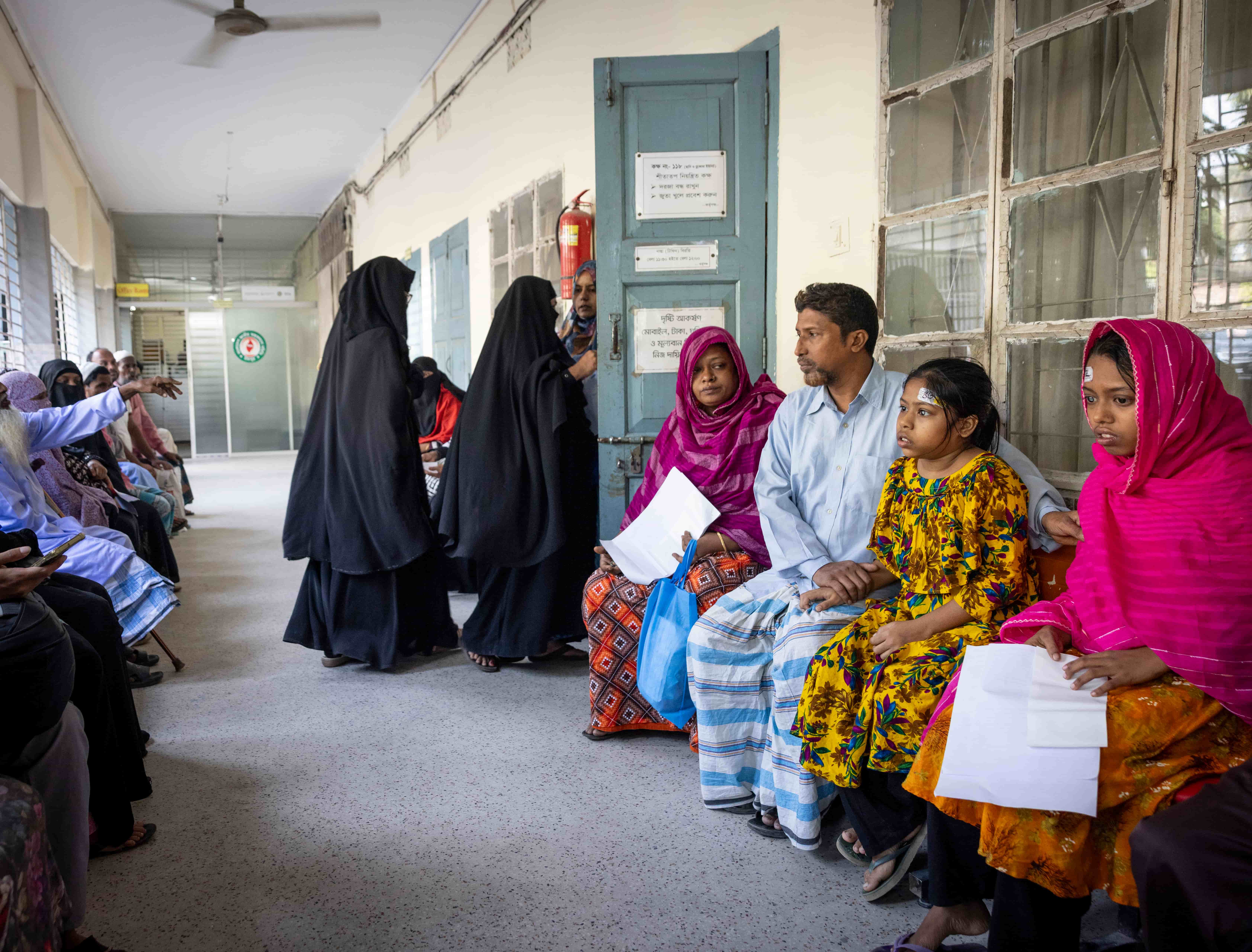 Sumaiya and Habiba sit quietly among other patients in a hospital hallway in Chandpur, Bangladesh, waiting to be screened. They sit close together on a bench, surrounded by families seeking eye care.