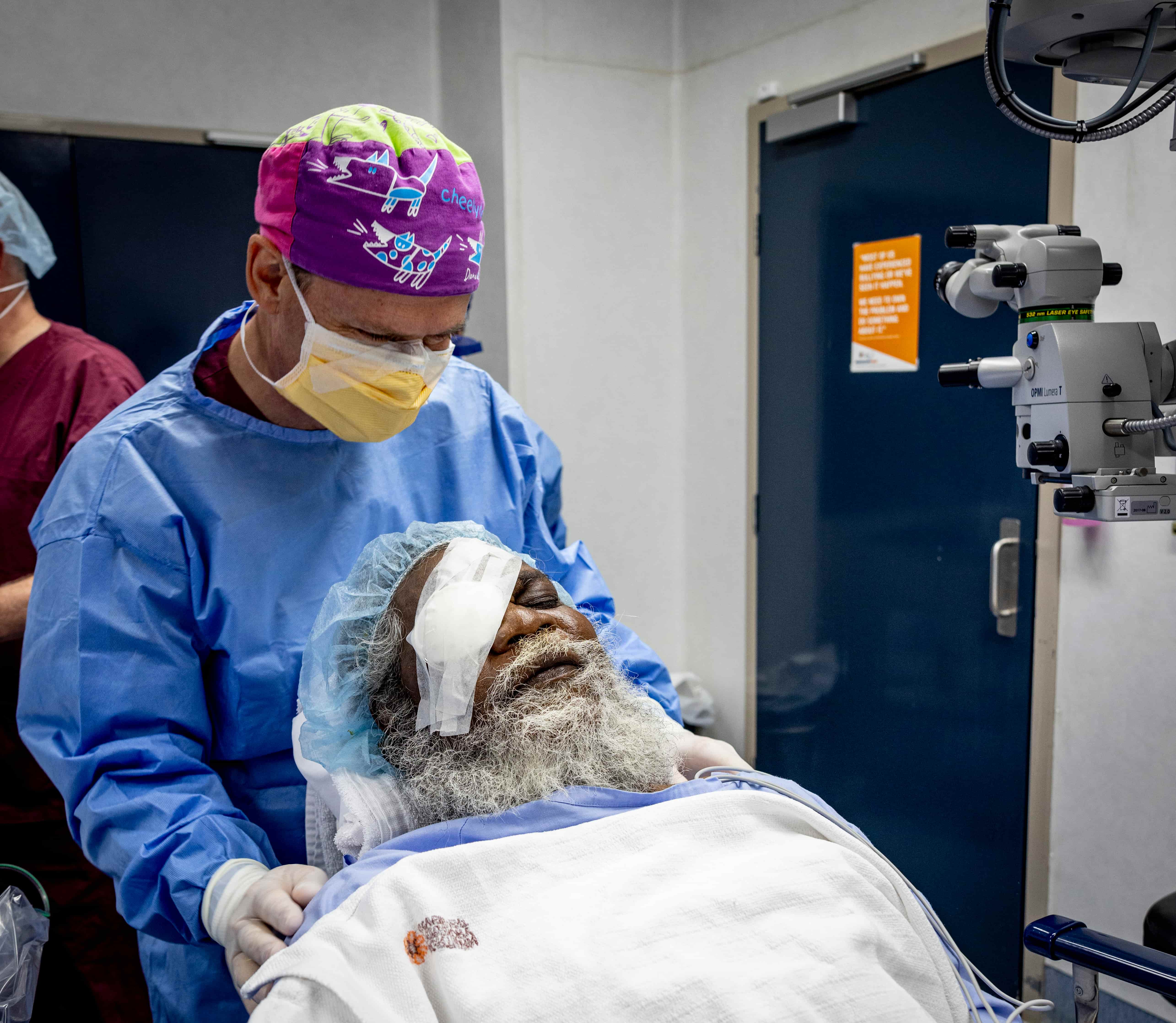 A photo of a doctor attending to a patient after eye surgery. The patient is an older man with a beard, and a patch is over one of his eyes. The doctor is standing over him, gently touching his shoulder.