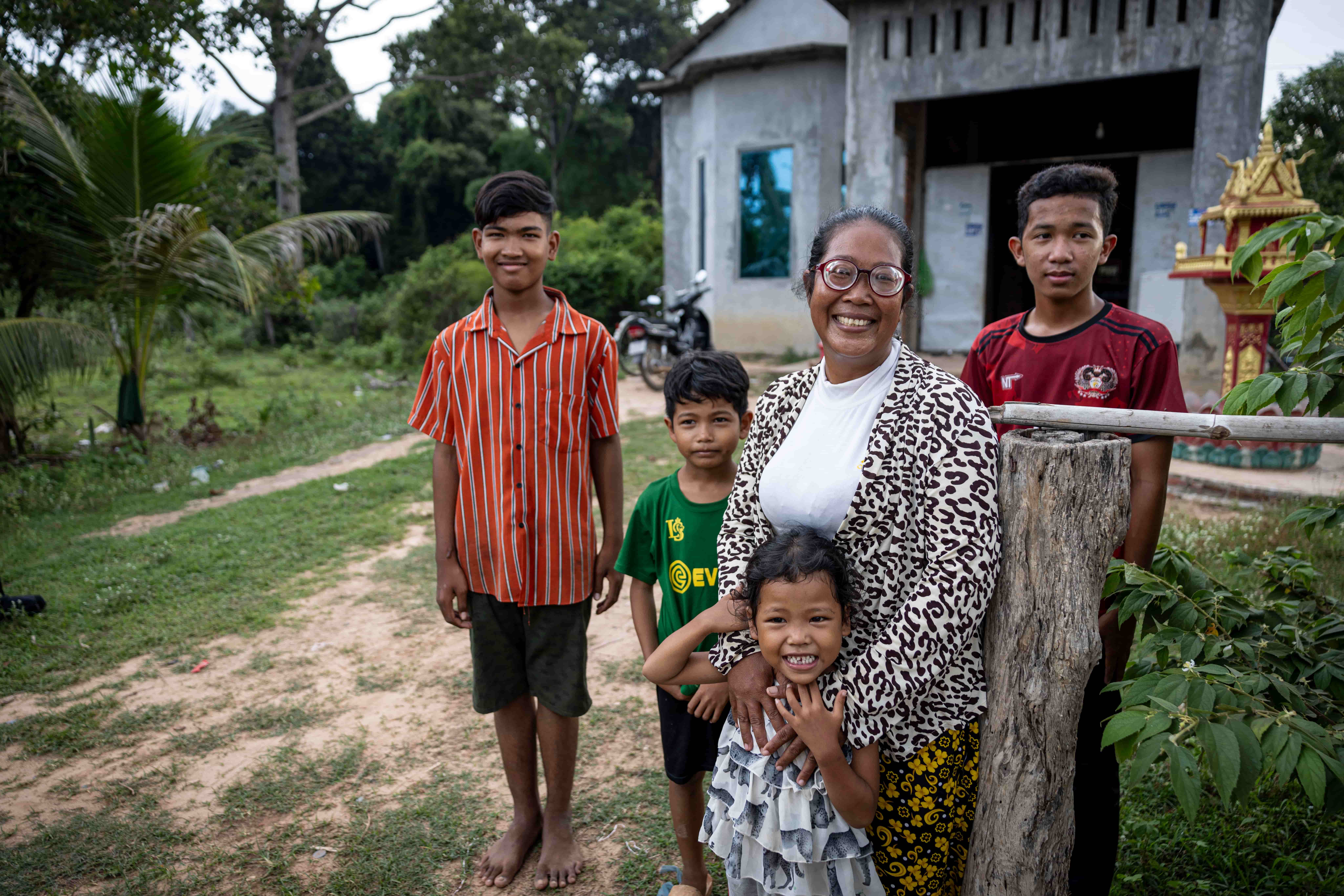 A photo of a woman with four children standing outside a house. All of them are smiling and looking at the camera. The setting appears to be a rural village.