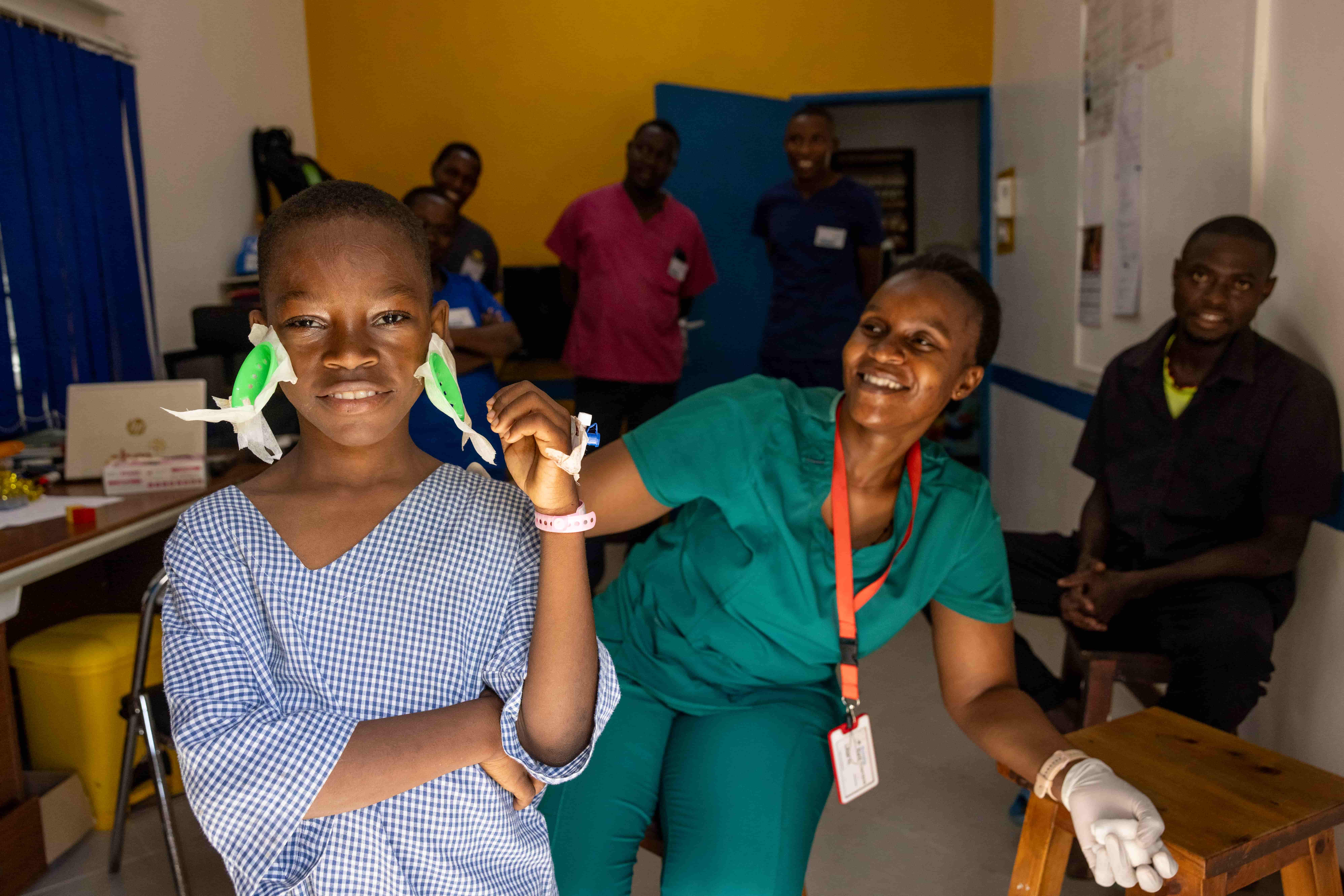 A photo shows a child in a hospital gown, smiling after undergoing cataract surgery. Post-operative eye patches are visible on their face. A healthcare worker is smiling and leaning down next to the child.