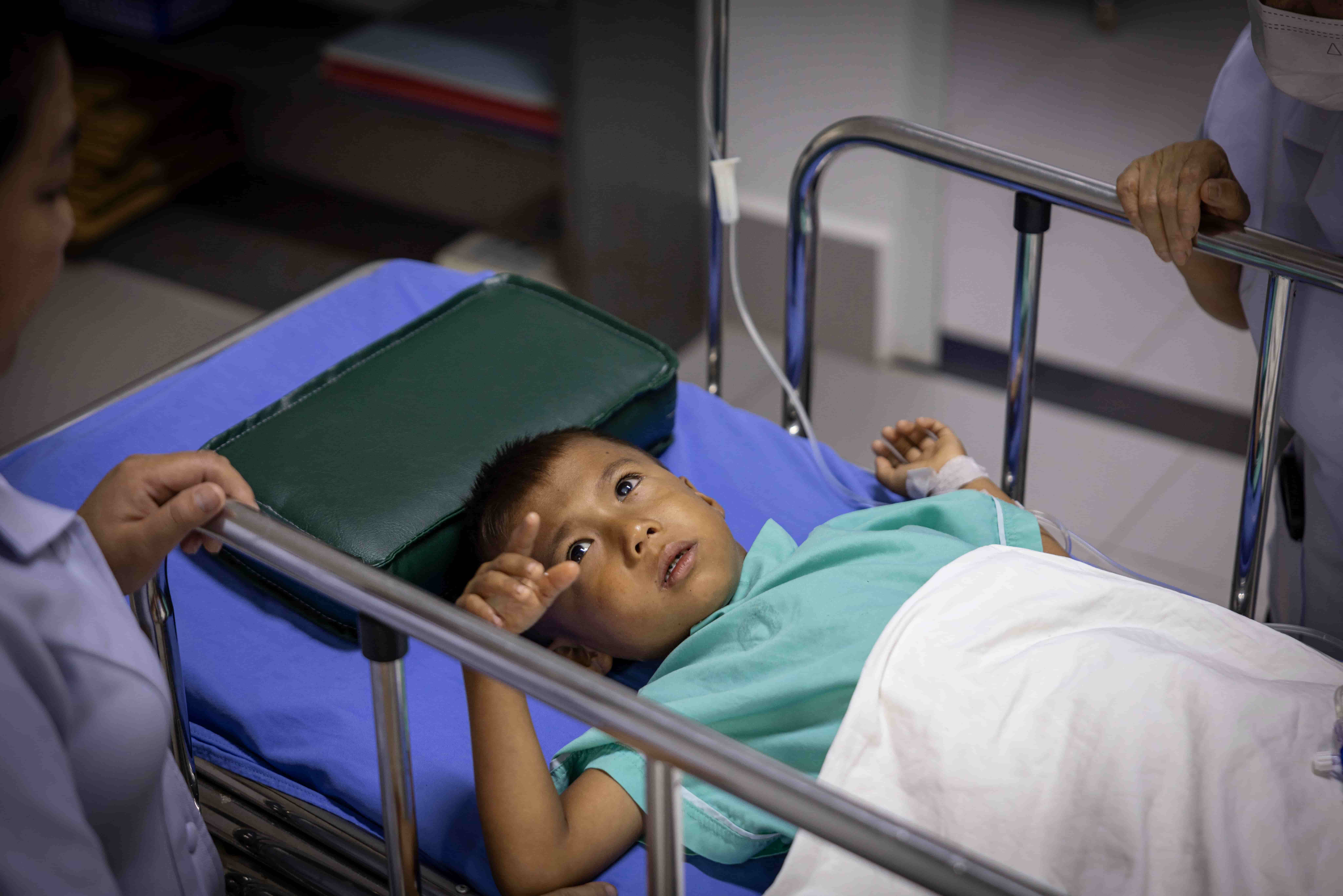 A photo of a young child lying on a hospital gurney. He looks at the camera with his hand slightly raised. The hands of two medical workers are on the sides of the gurney.