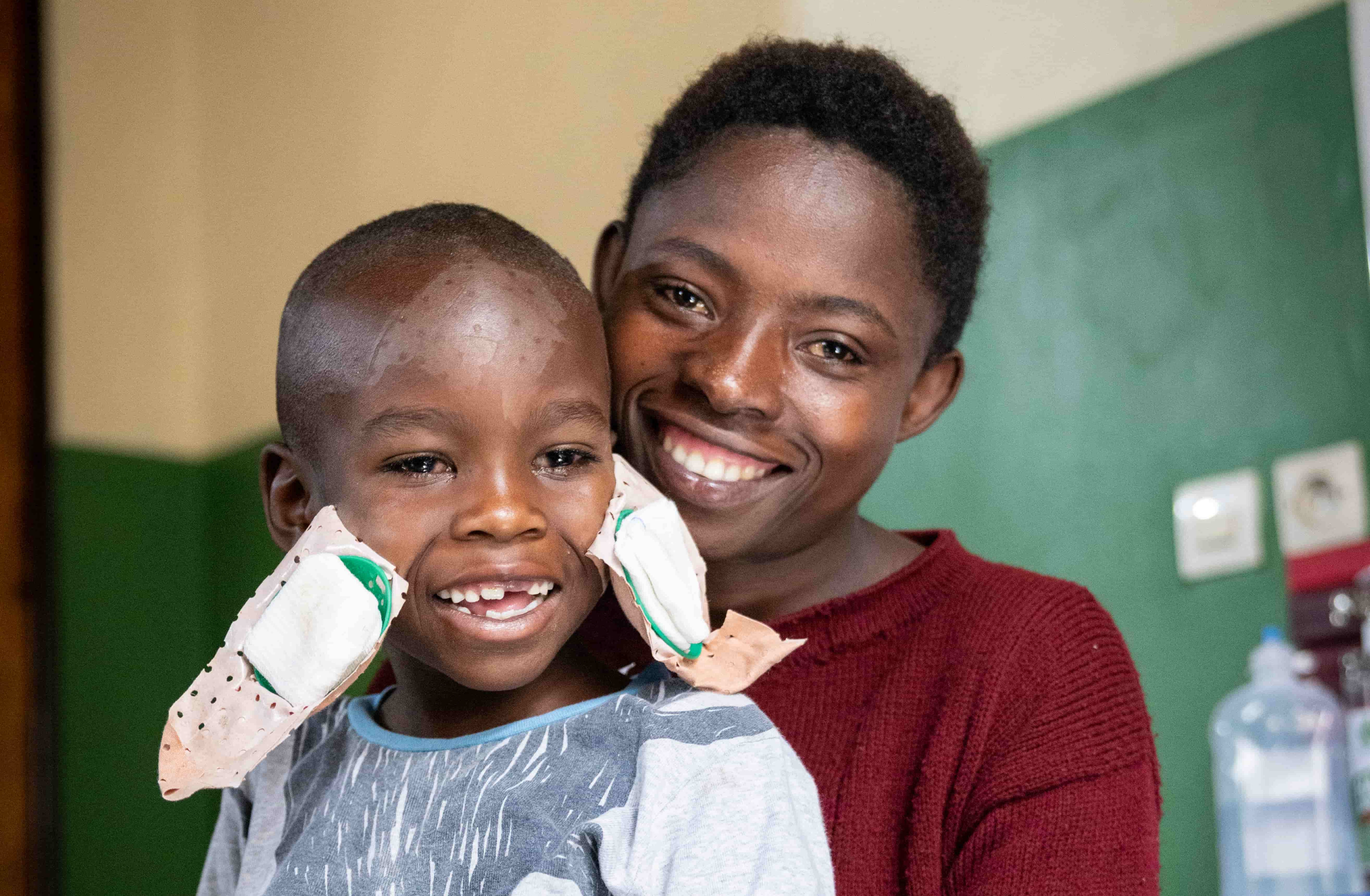 A smiling child with eye bandages sits in front of his smiling mother in a red sweater.