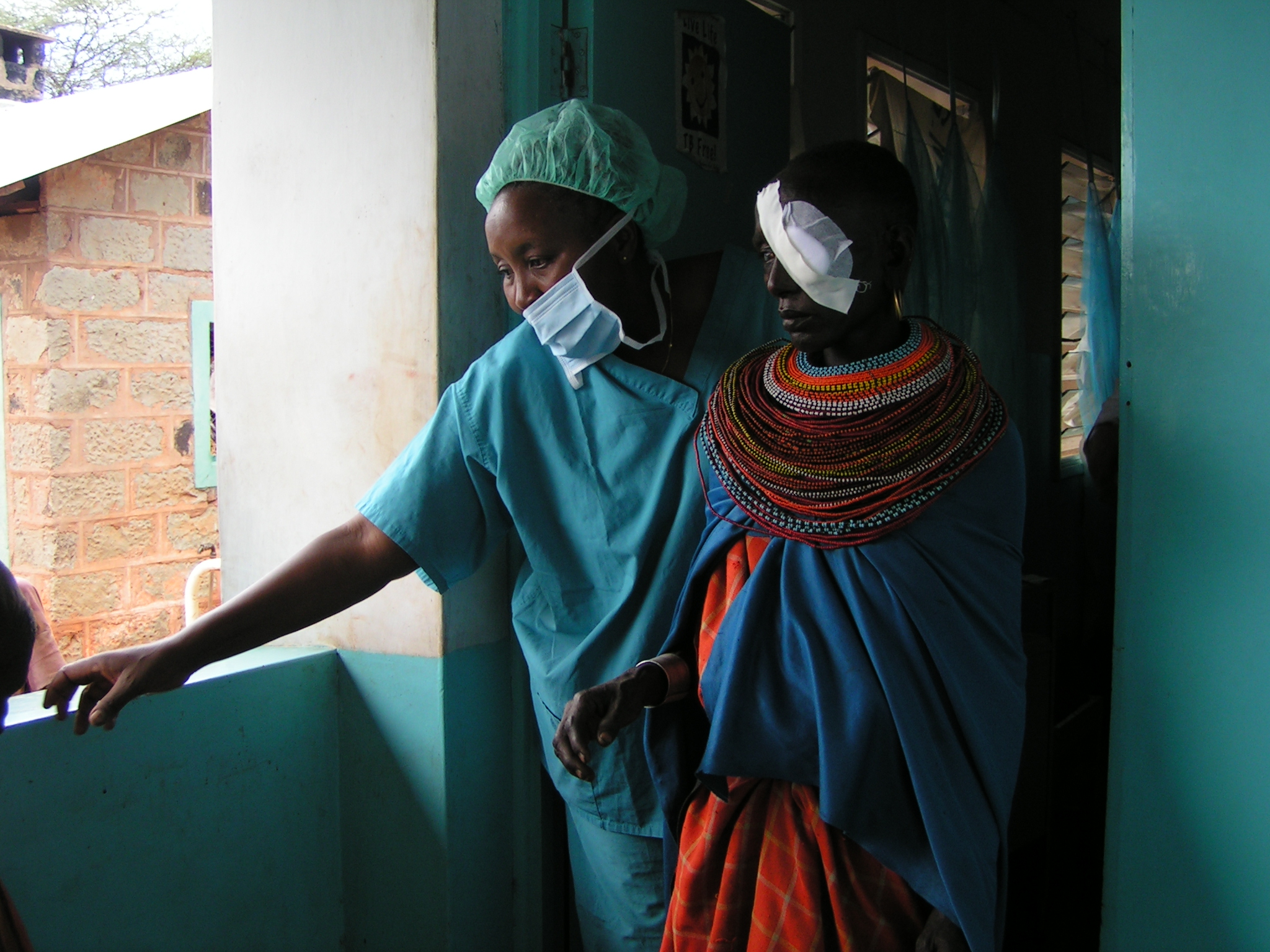 Rose being led by the surgeon after being operated on. She has a patch over her eye and traditional Kenyan beads around her neck and gold hoops in her ears.